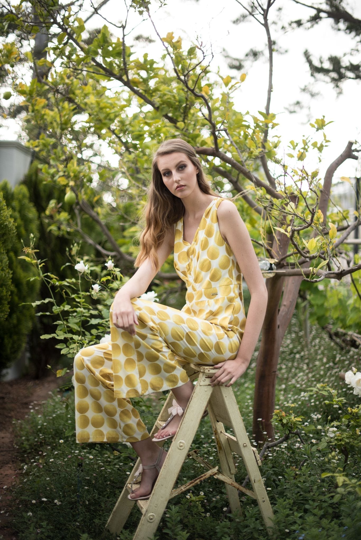 Model in yellow polka dot jumpsuit sitting on vintage ladder in garden